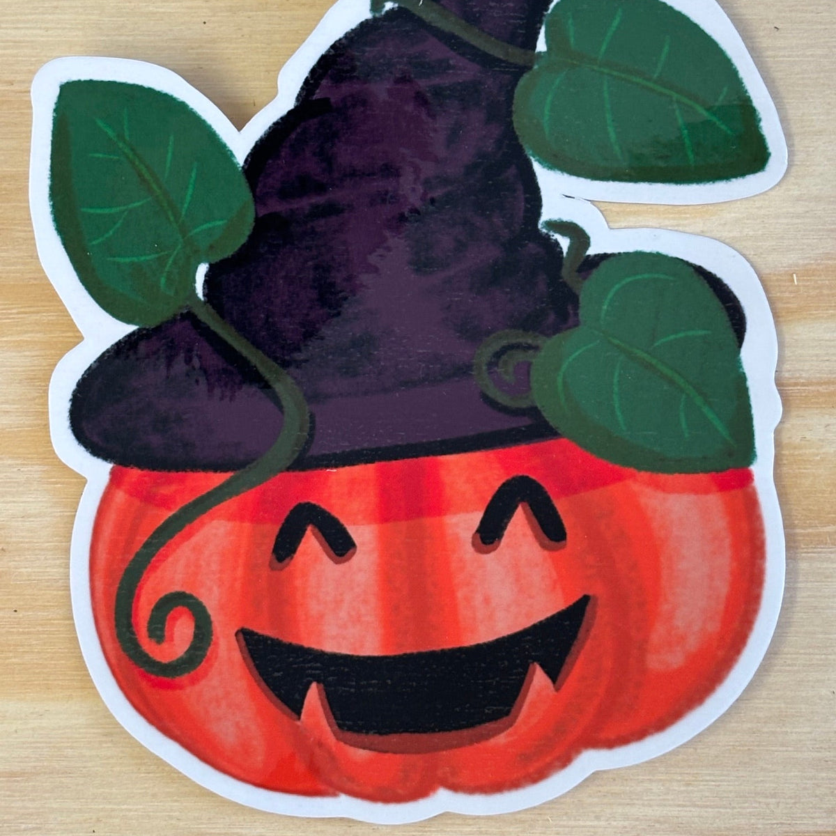 A die-cut sticker of a smiling orange pumpkin wearing a dark purple witch hat with green leaves, placed on a light wood background.