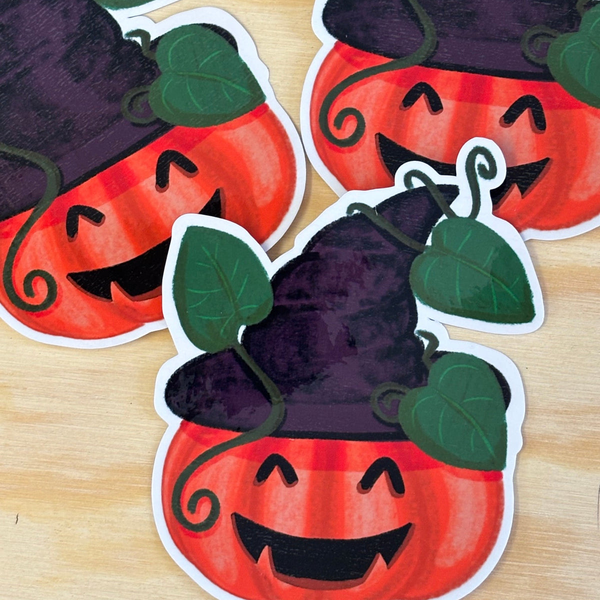 Three Halloween pumpkin stickers with carved faces, green leaves, and purple witch hats arranged on a light wood surface.
