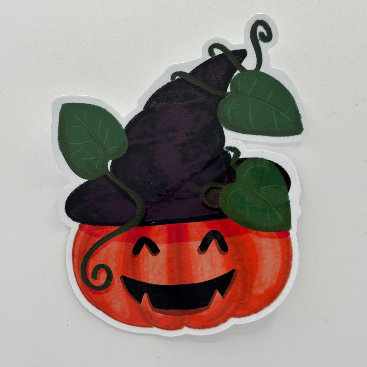 A die-cut sticker featuring a cute Halloween design of a smiling orange pumpkin wearing a dark purple witch hat with green leaves, shown on a plain light gray background.