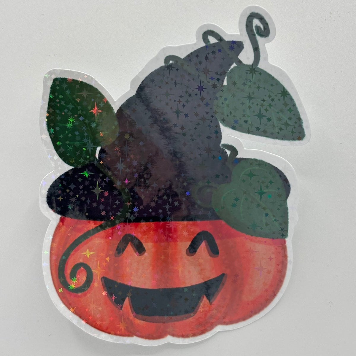 A die-cut sticker featuring a smiling orange jack-o'-lantern with a black witch hat and green leaves, filled with glitter, on a light gray background.