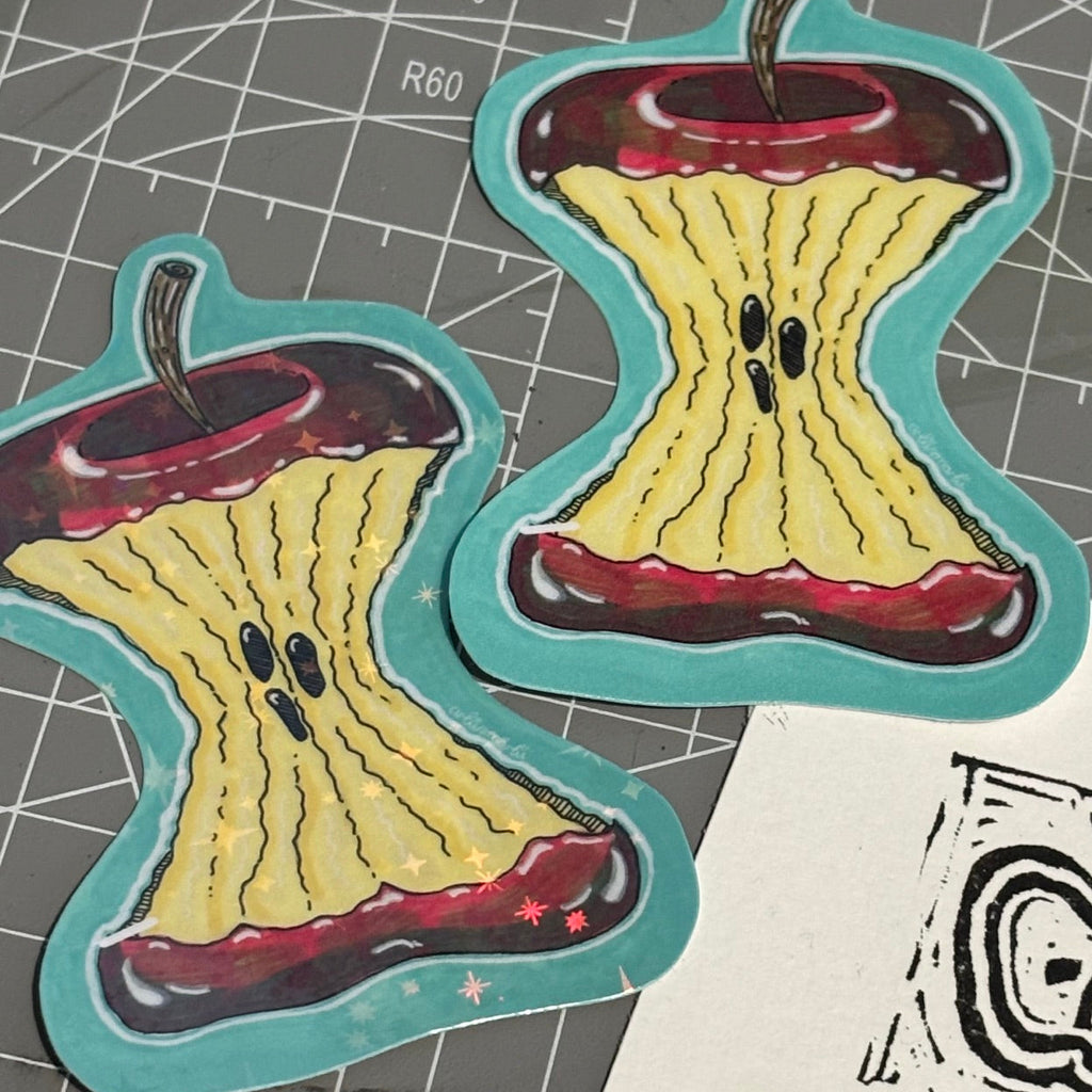 Two die-cut stickers shaped like stylized apple cores with yellow centers, red ends, and teal borders, placed on a gray cutting mat.