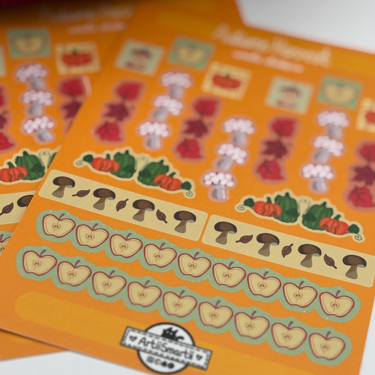 A close-up of two orange sticker sheets featuring autumn-themed illustrations, including pumpkins, mushrooms, acorns, and flowers, with a white background.