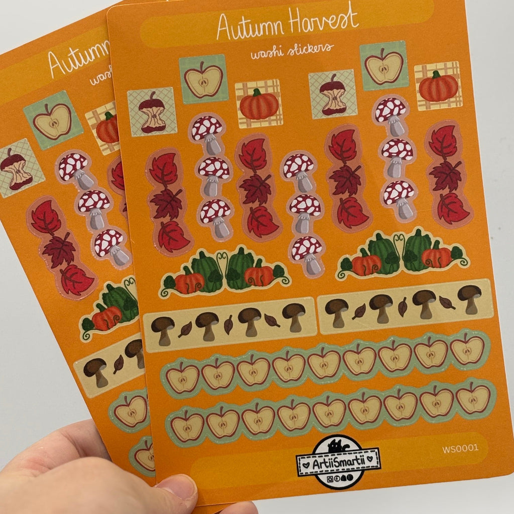 A hand holds two orange planner stickers labeled 'Autumn Harvest' against a plain light background. The stickers feature illustrated autumn-themed elements including pumpkins, mushrooms, leaves, and apples.