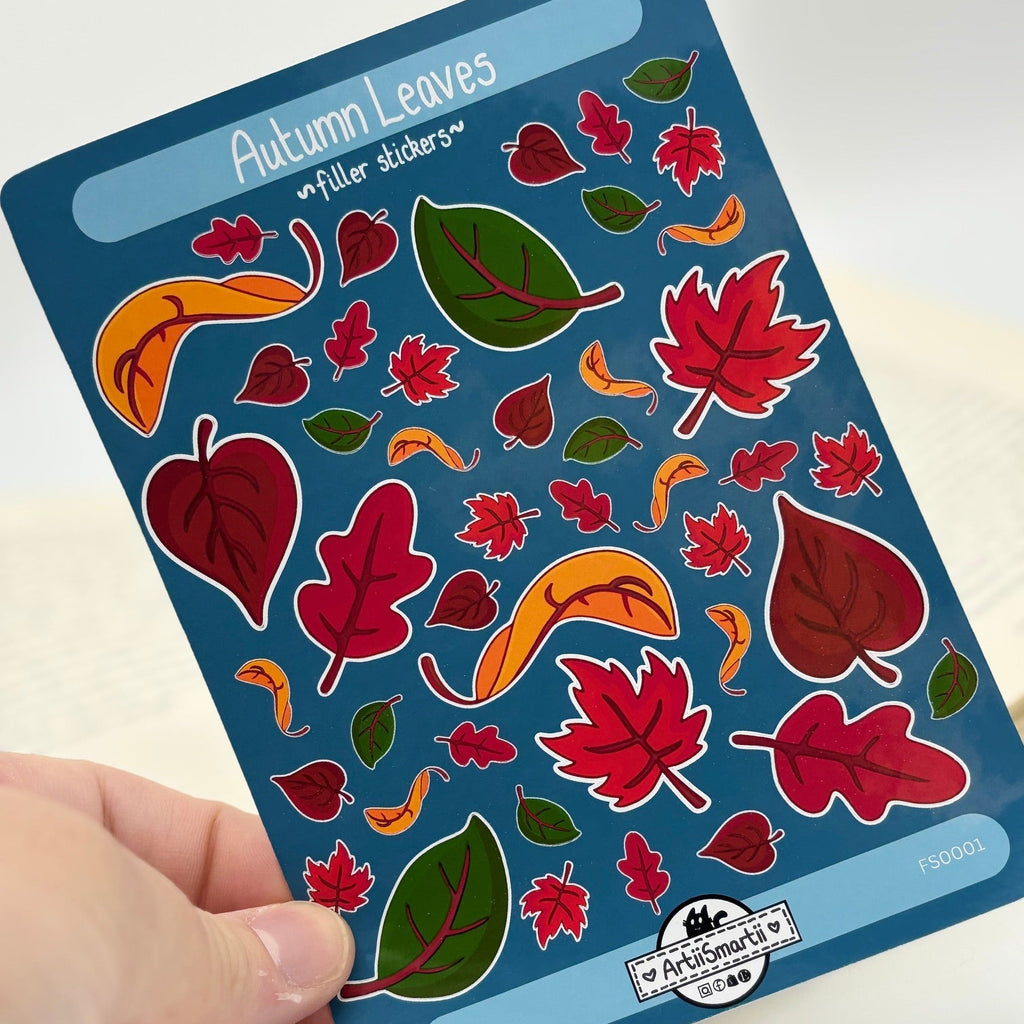 A hand holds a blue sticker sheet labeled 'Autumn Leaves' featuring colorful leaf and maple leaf stickers in red, green, orange, and brown, with a logo for Art of Crafts' Way at the bottom.