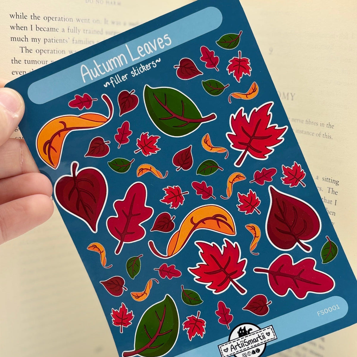 A hand holds a blue sticker sheet labeled 'Autumn Leaves' with colorful illustrated leaves in red, green, and orange, with an open book in the background.