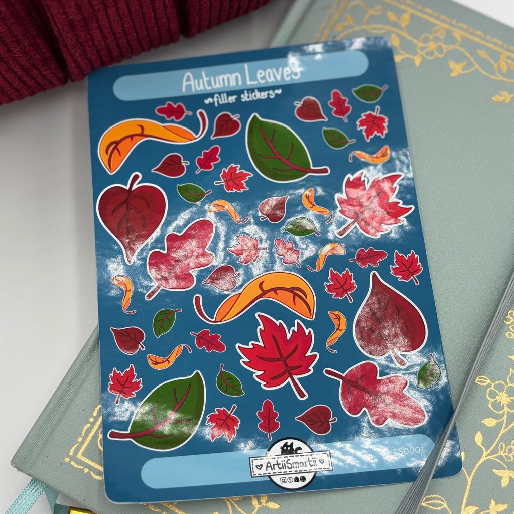 A sheet of 'Autumn Leaves' paper stickers with colorful red, orange, and green leaves on a blue backing, styled on top of a green patterned notebook with a burgundy knit sweater in the corner.