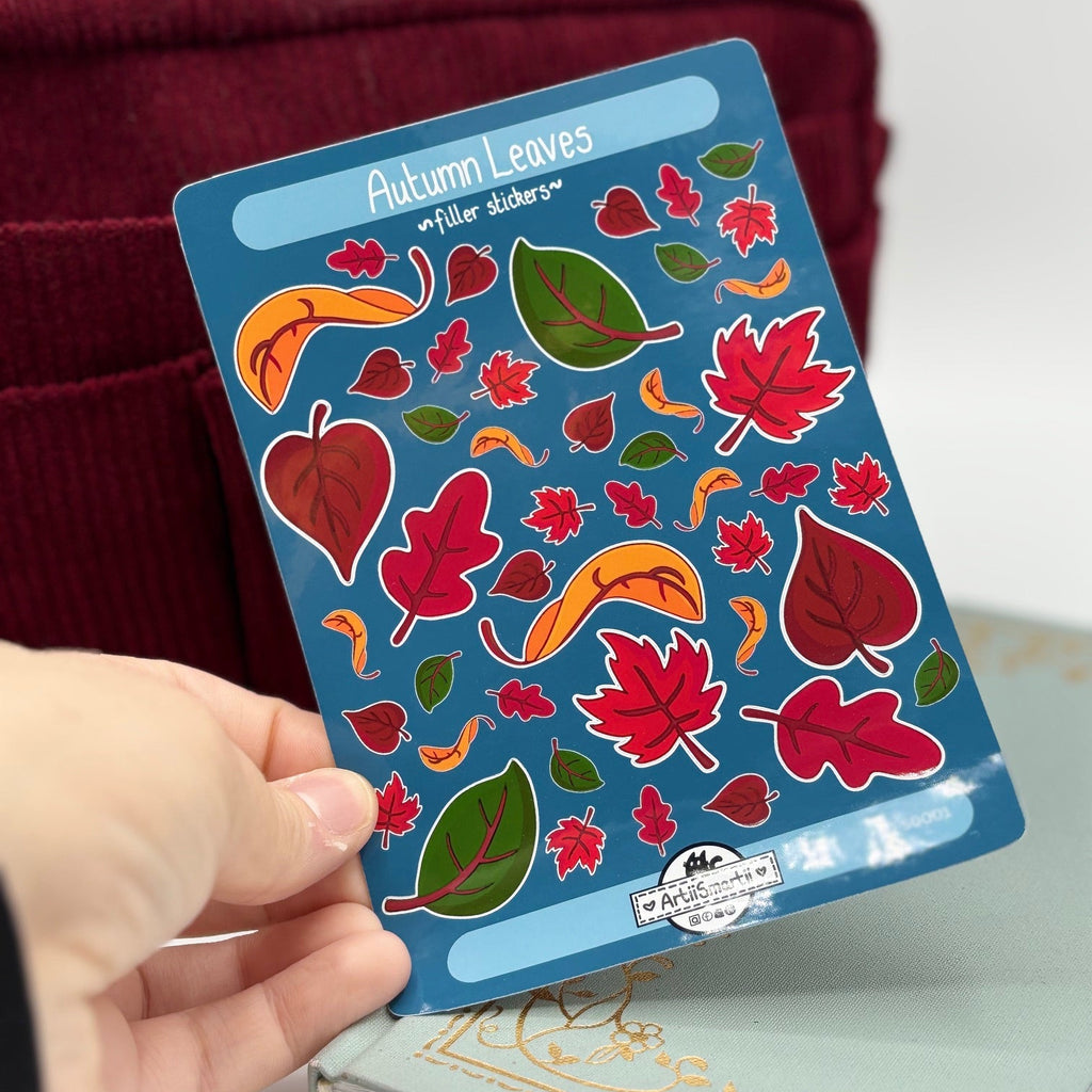 A hand holds a blue sticker sheet labeled 'Autumn Leaves Sticker Sheets' featuring colorful red, orange, green, and yellow leaf illustrations. The background includes a dark red corduroy bag and a light-colored notebook.