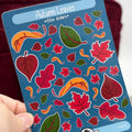 A hand holds a blue sticker sheet labeled 'Autumn Leaves filler stickers' featuring colorful illustrated leaves in red, orange, and green. The brand name 'Artiismartii' is visible at the bottom of the sheet.