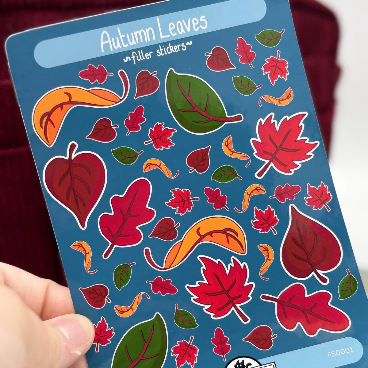 A hand holds a blue sticker sheet labeled 'Autumn Leaves filler stickers' featuring colorful illustrated leaves in red, orange, and green. The brand name 'Artiismartii' is visible at the bottom of the sheet.
