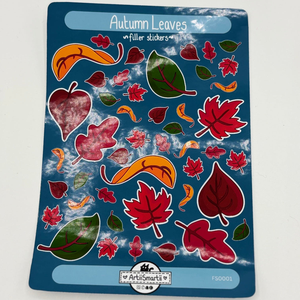 A sheet of 'Autumn Leaves' fuller stickers by Artiismartii, featuring various red, orange, and green leaf designs on a dark blue backing card.