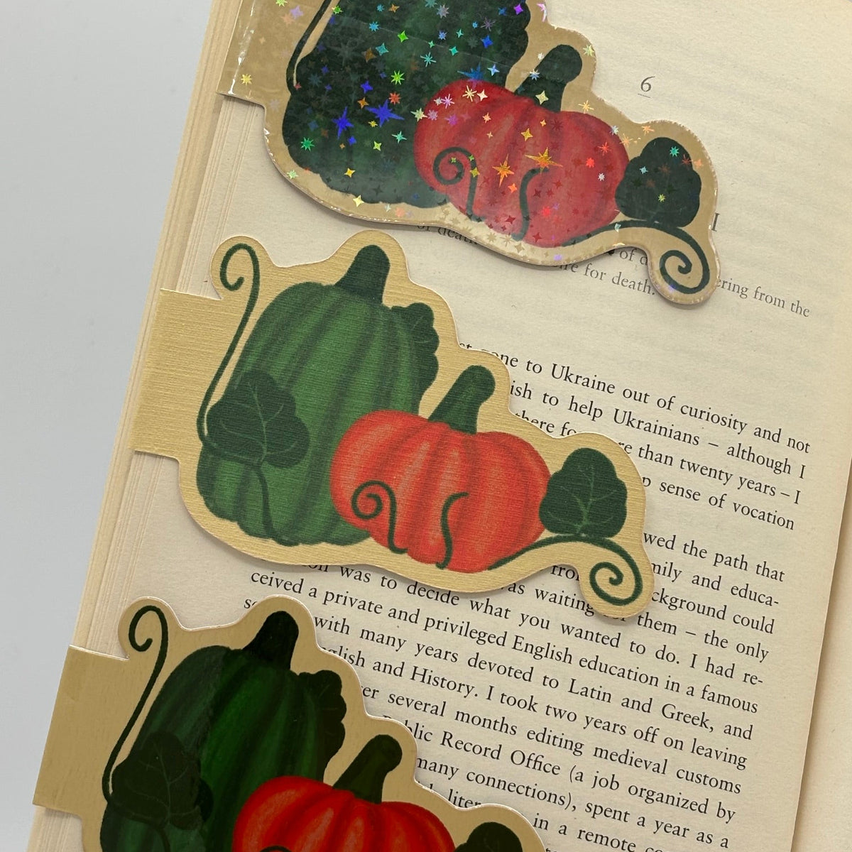 Three autumn-themed wooden bookmarks shaped like pumpkins and gourds are arranged on an open book. The bookmarks feature natural wood edges with black scroll details and are decorated with red, orange, and green painted motifs, including one with a glittery dark green top.