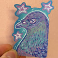 A hand holding a die-cut sticker featuring a stylized purple and teal pigeon head with pink and white stars on a peach-colored background.