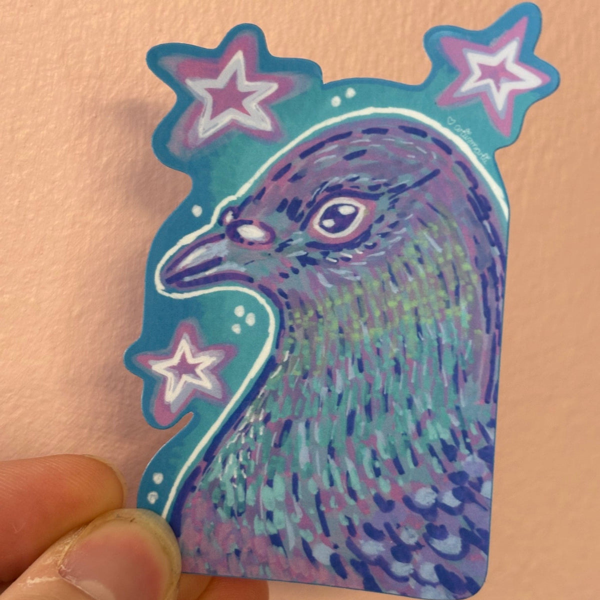 A hand holding a die-cut sticker featuring a stylized purple and teal pigeon head with pink and white stars on a peach-colored background.