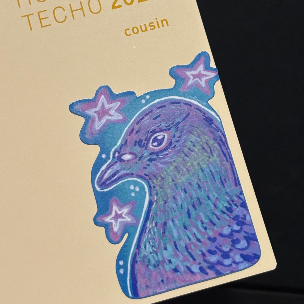 A close-up of a Hobonichi Tech O 2027 card featuring a stylized purple and blue bird illustration with pink stars on a cream-colored card, set against a dark background.