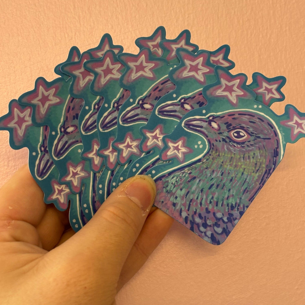 A hand holding a fan of die-cut stickers featuring a stylized pigeon and stars in teal, purple, and pink, set against a plain peach-colored wall.